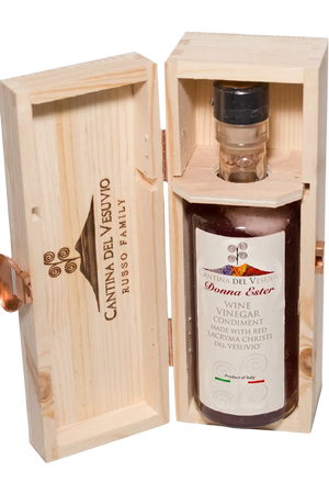 A bottle of Vessuvio wine vinegar presented in a light natural wood gift box with the lid leaning against the base. The bottle has a white label and a black cap, and the wooden box is engraved with the brand name and a grape logo.